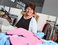 Eine Frau mittleren Alters mit rot gefärbten Haaren steht hinter der Kassa des carla Shops in Altach. Sie trägt eine Brille, ein schwarzes Oberteil und darüber eine graue Stoffjacke. 