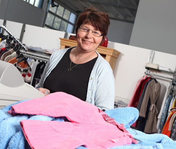 Eine Frau mittleren Alters mit rot gefärbten Haaren steht hinter der Kassa des carla Shops in Altach. Sie trägt eine Brille, ein schwarzes Oberteil und darüber eine graue Stoffjacke. 