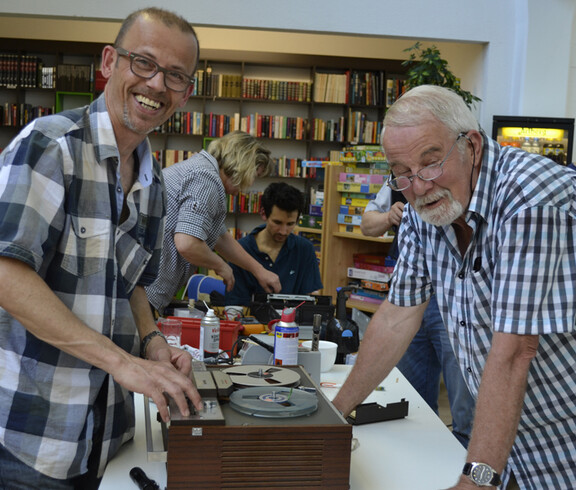 Ein älterer Herr und Mann mittleren Alters, die beide karierte Hemden tragen, reparieren ein altes Aufnahmegerät, das auf einem weißen Tisch steht. Die Stimmung ist entspannt und beide lachen. 