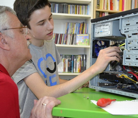 Generationenaustausch: Ein junger Bursche mit grauem t-Shirt und ein älterer Mann mit rotem Polo reparieren einen Computer. Die jeweiligen Hardwareteile sind dabei gut ersichtlich. Der PC steht auf einem grünen Tisch. 