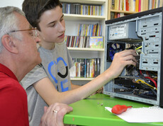 Generationenaustausch: Ein junger Bursche mit grauem t-Shirt und ein älterer Mann mit rotem Polo reparieren einen Computer. Die jeweiligen Hardwareteile sind dabei gut ersichtlich. Der PC steht auf einem grünen Tisch. 