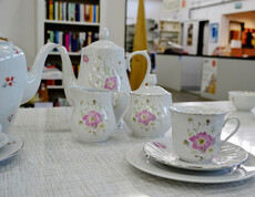 Weißes Kaffeegeschirr mit einer rosa Blume steht auf einem weißen Tisch. 