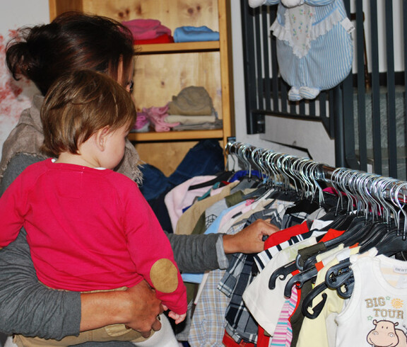 Eine Mutter mit Kind ist auf der Suche nach dem passendem Gewand. Sie stehen vor einem Kleiderständer. Die Mutter hat dabei das kleine Kind auf dem Arm. 