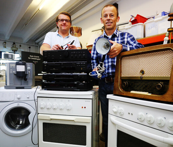 Zwei Männer mittleren Alters stehen hinter defekten Elektogeräten. Das sind unter anderem Lautsprechanlagen und weißen Waschmaschienen. 