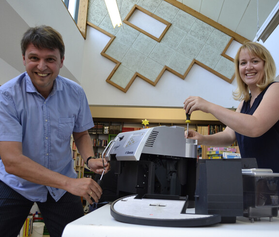 Zwei erwachsene Menschen mittleren Alters, ein Mann und eine Frau, reparieren eine Kaffeemaschine. Der Mann trägt ein blaues Hemd und die Frau ein dunkelblaues Kleid. 